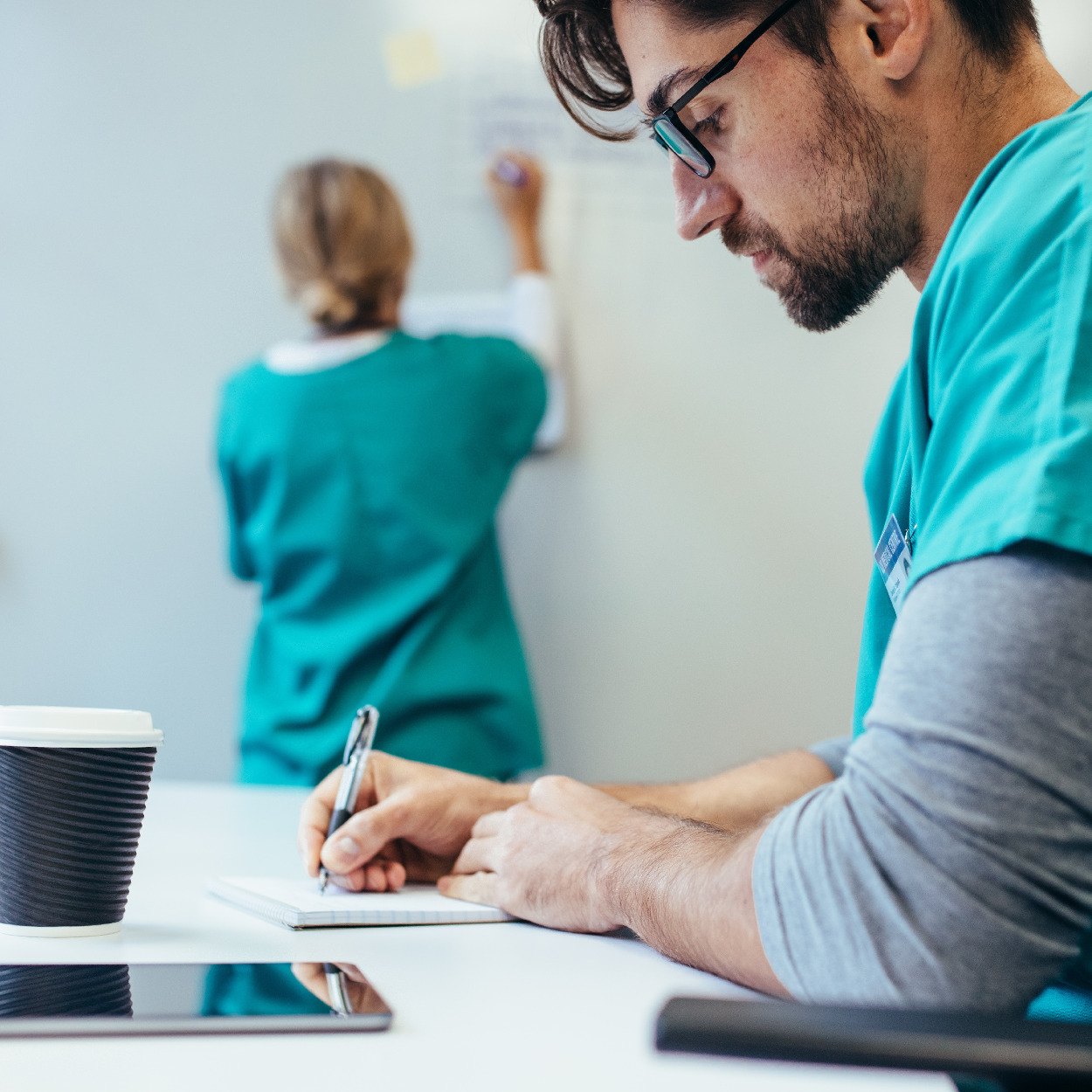 Male nurse in green scrubs staring down while he's writing notes on a pad of paper